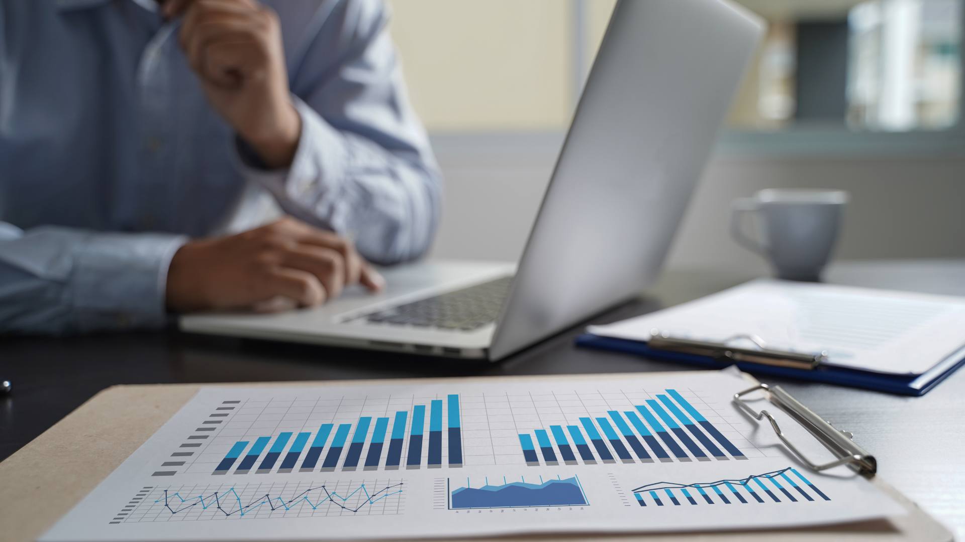 Profissional analisando gráficos de desempenho financeiro em planilhas e laptop, representando o uso de dados e tecnologia para previsão de inadimplência em empresas.